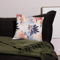 Decorative pillow on a dark sofa draped with an olive green blanket, featuring a winter bloom floral design in soft pink, blue, and neutral tones.