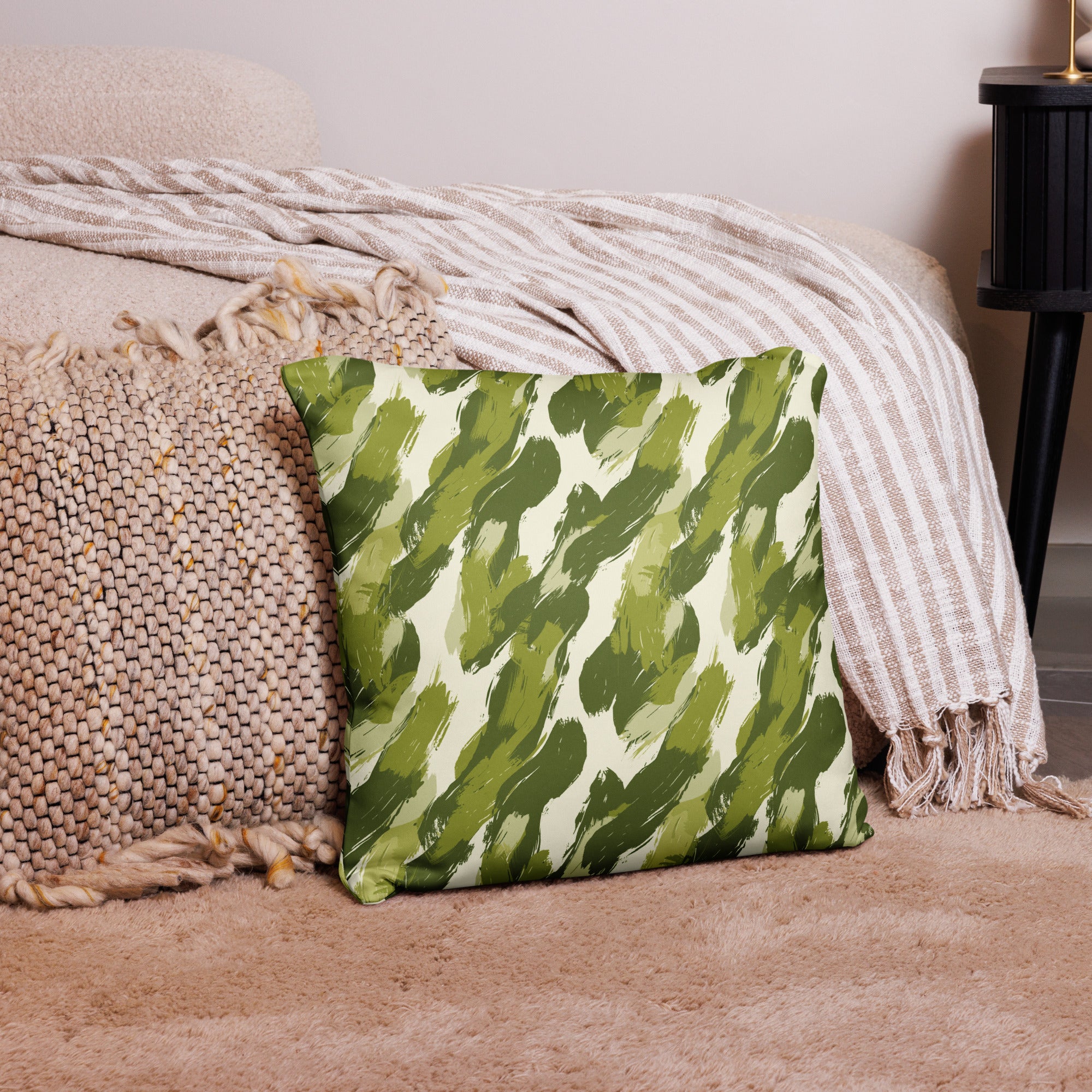 Plush pillow with green brushstroke jungle design on beige fabric, placed on a cozy textured rug with woven throw blankets in the background.