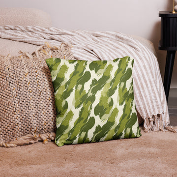 Plush pillow with green brushstroke jungle design on beige fabric, placed on a cozy textured rug with woven throw blankets in the background.