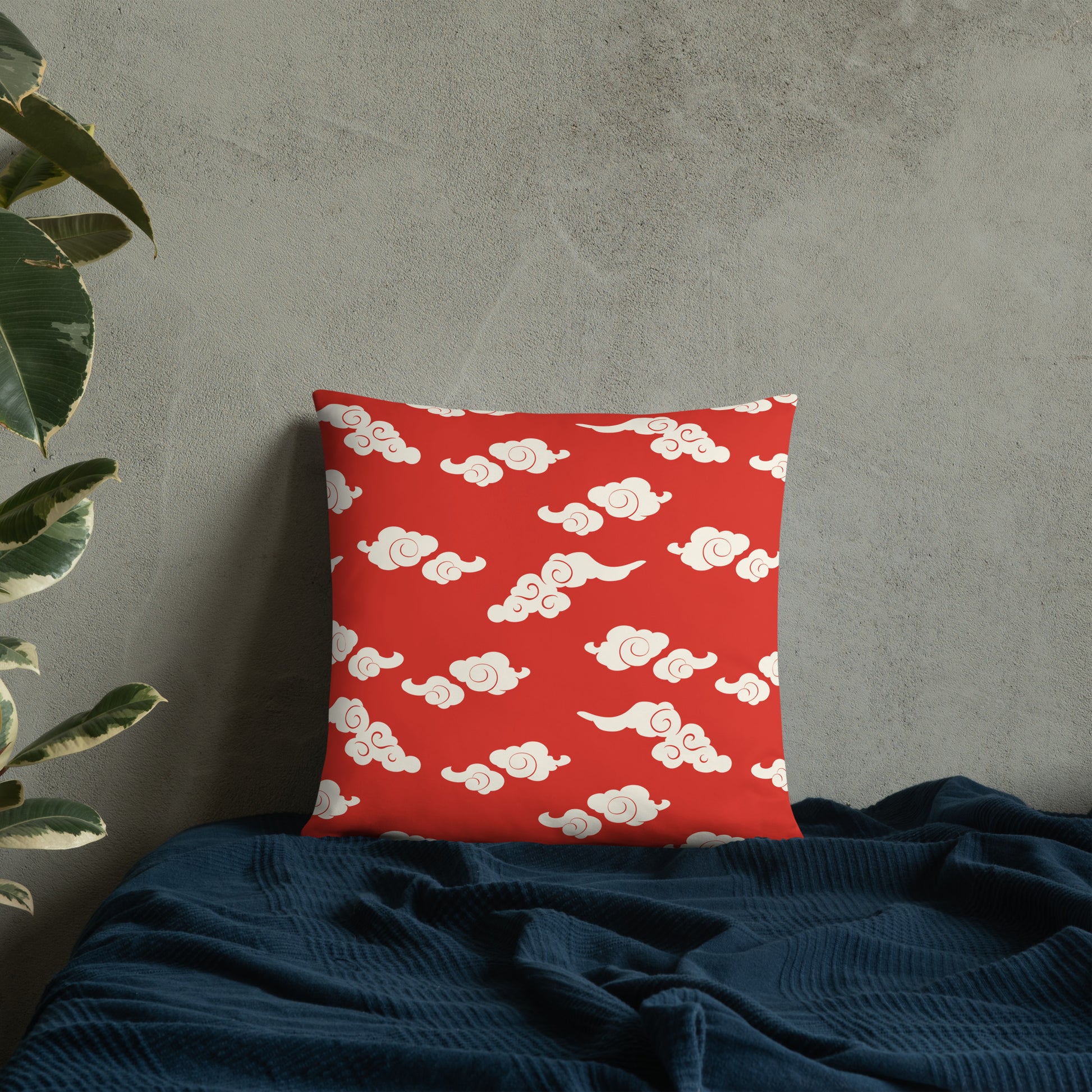 Red accent pillow with whimsical white cloud pattern on a grey textured wall background, nestled on a dark blue blanket.