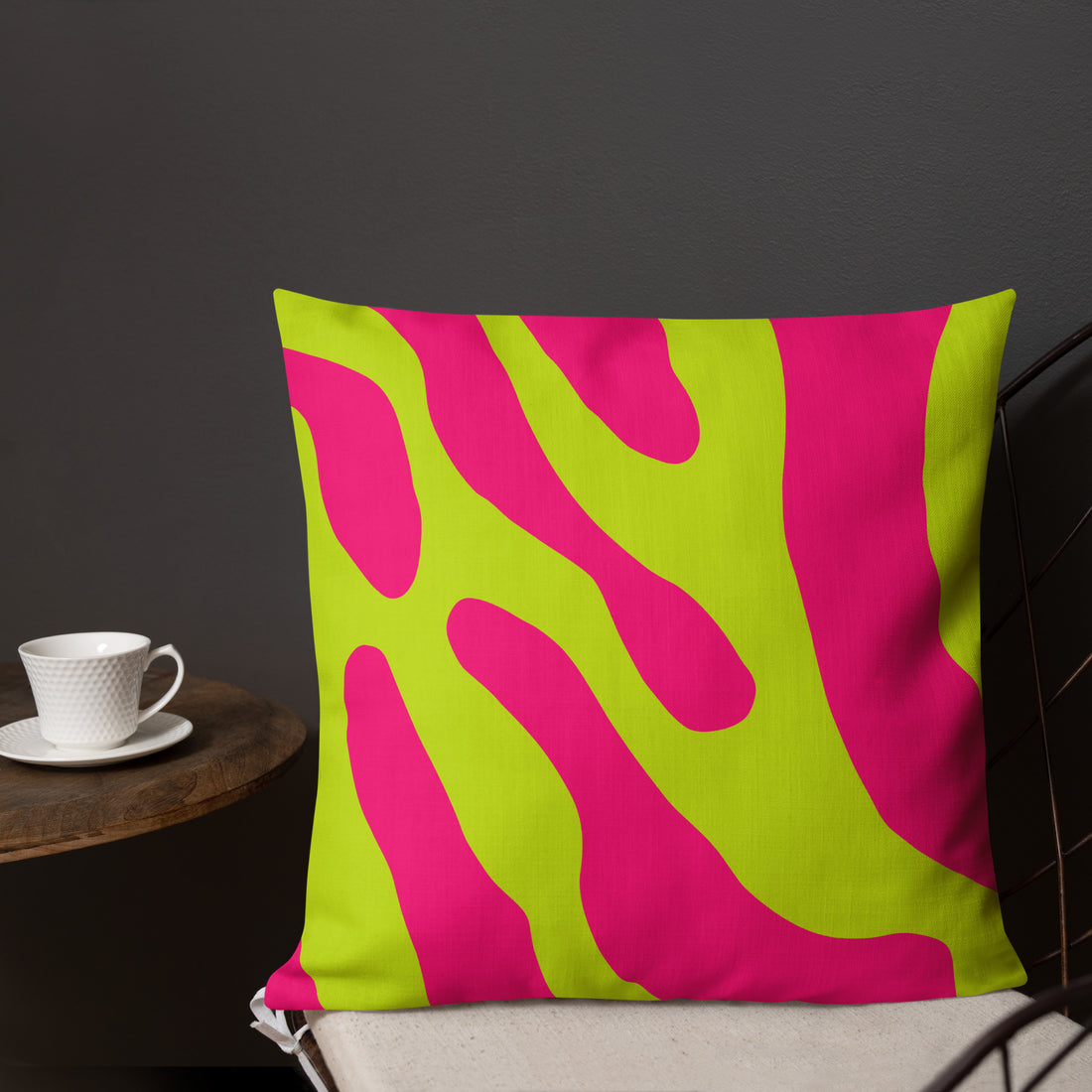 Vibrant abstract wave pattern pillow in pink and lime green colors on a chair with a cup nearby.