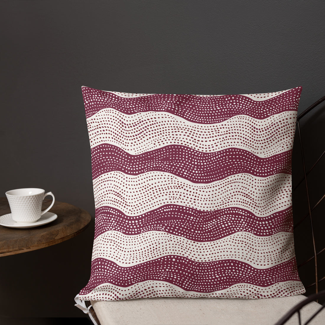 Burgundy knitted pillow with white wave patterns, cozy linen feel, placed on a chair next to a cup.