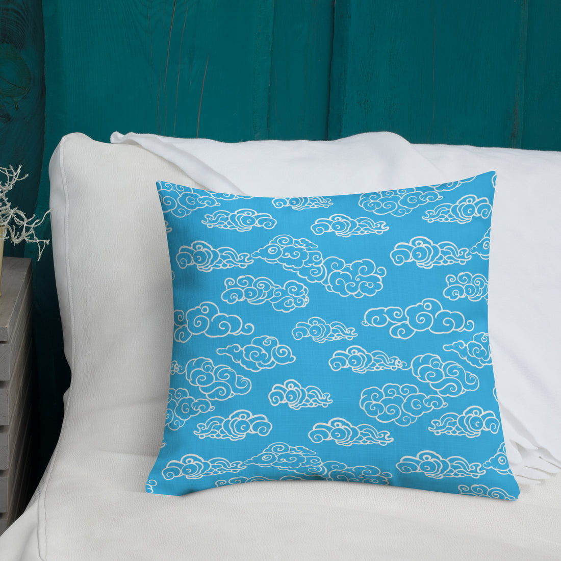 Azure blue pillow with white cloud patterns, knitted design, on a dark blue bedspread next to a plant.