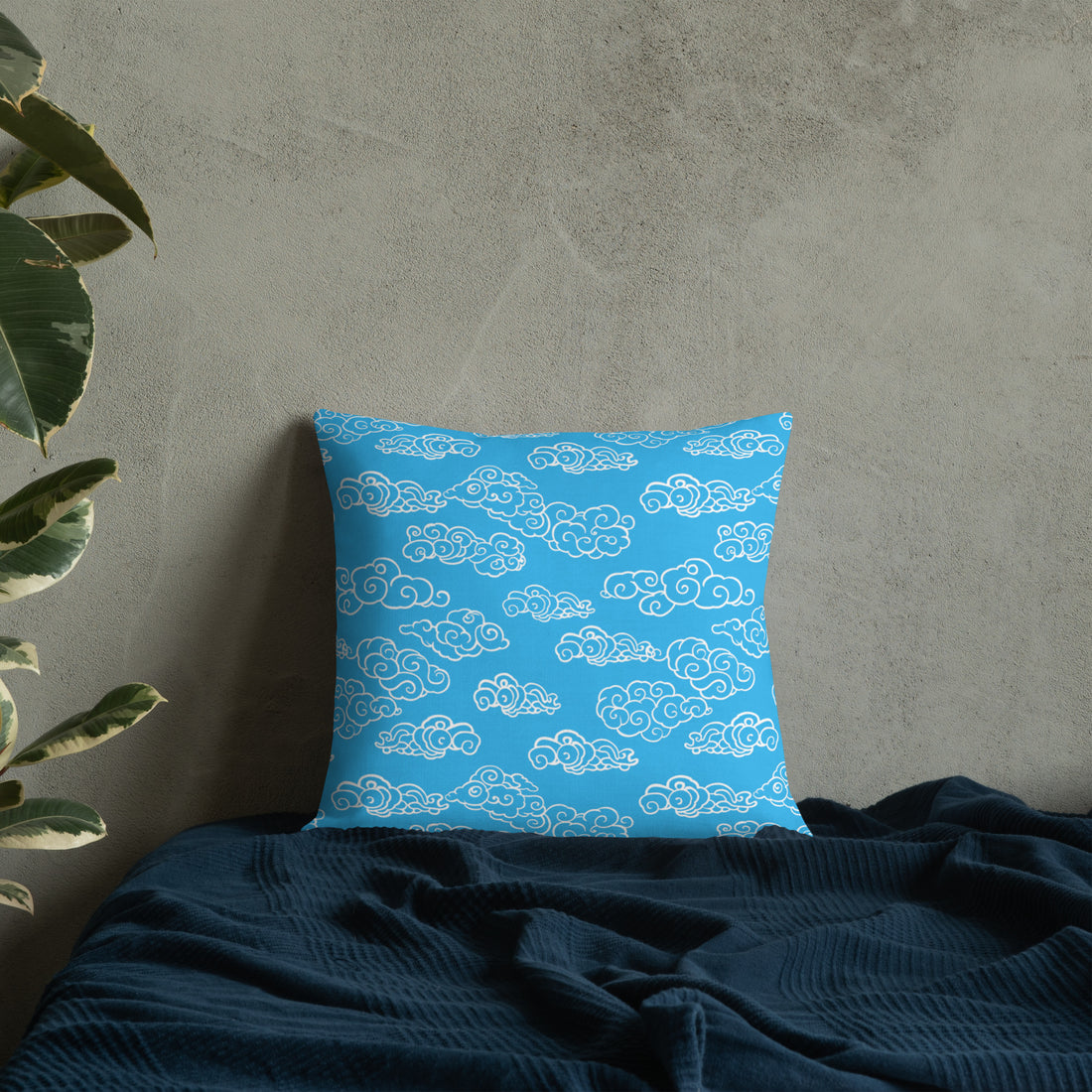 Azure blue pillow with white cloud patterns, knitted design, on a dark blue bedspread next to a plant.