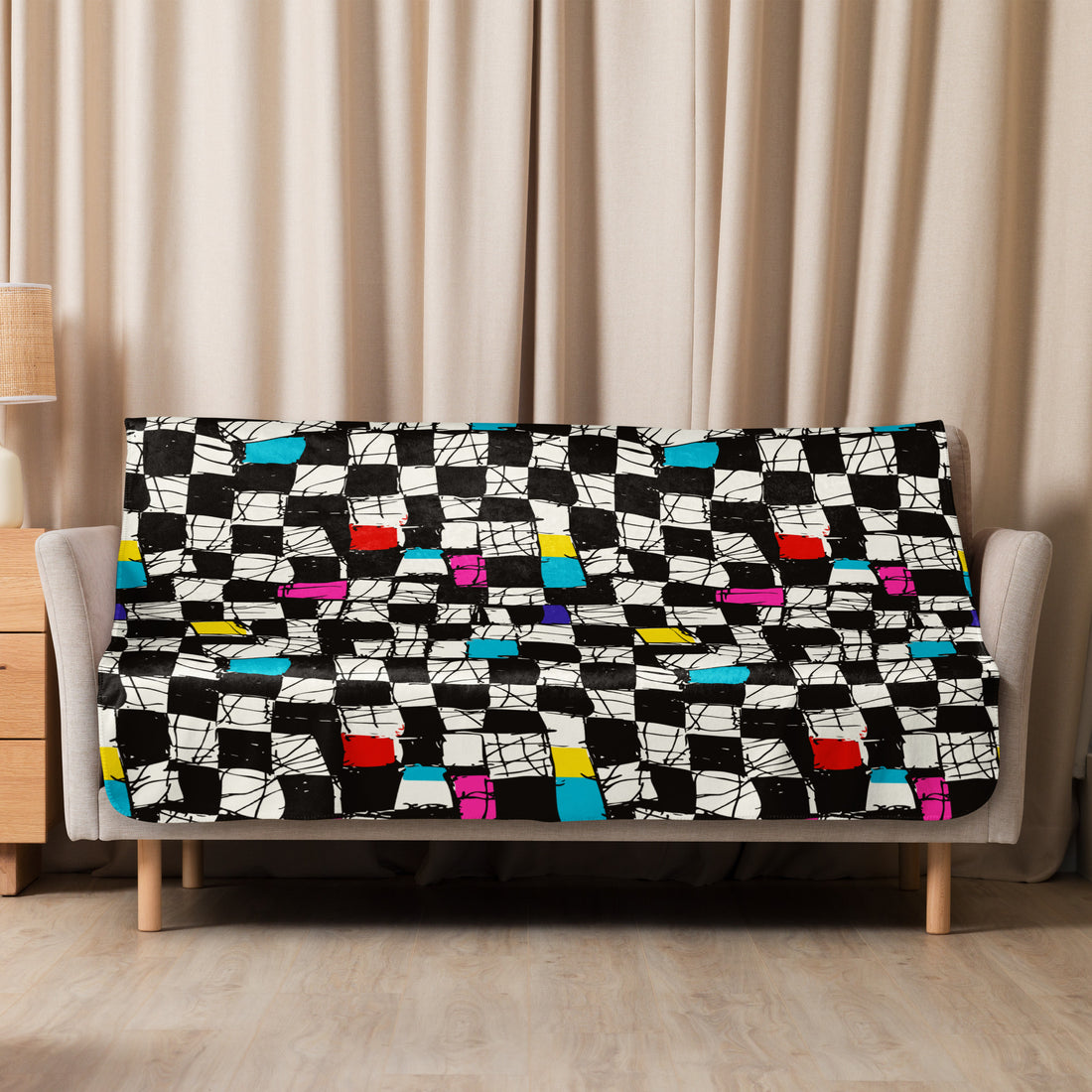 A sherpa blanket featuring a vibrant checkered design with splashes of red, blue, yellow, pink, and turquoise on a black and white background, draped over a couch.