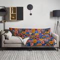 Knitted throw blanket with vibrant autumn leaves design in orange, blue, and red hues draped over a sofa.